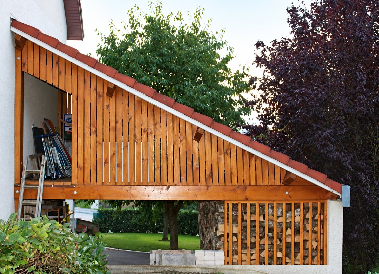 Un appui-toit en bois à claire-voie et toiture de tuiles rouges, adossé à une maison, avec du bois de chauffage empilé en dessous.