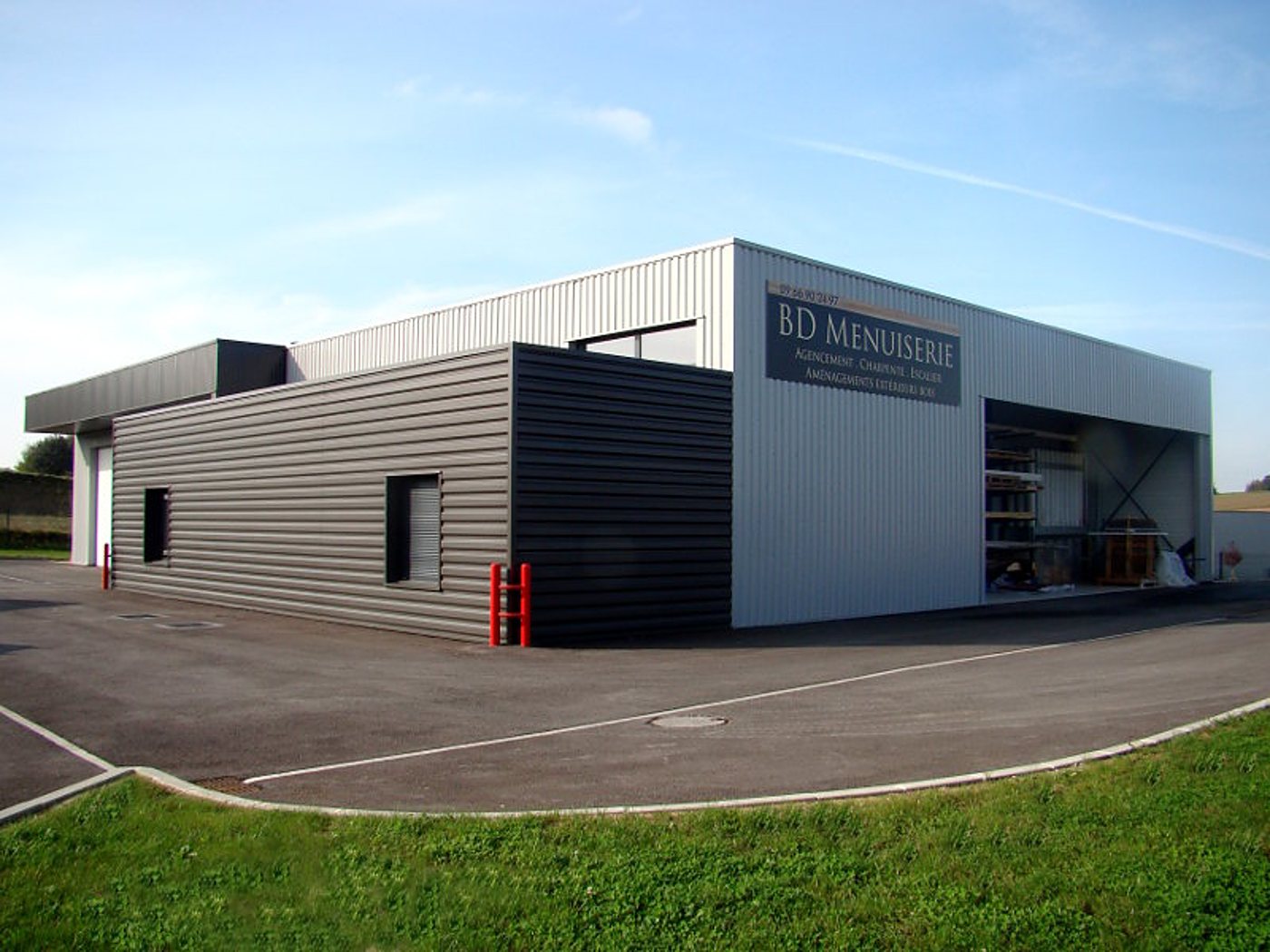 Façade d'un atelier à bardage métallique gris portant l'enseigne BD Menuiserie, avec un parking goudronné et une pelouse au premier plan.