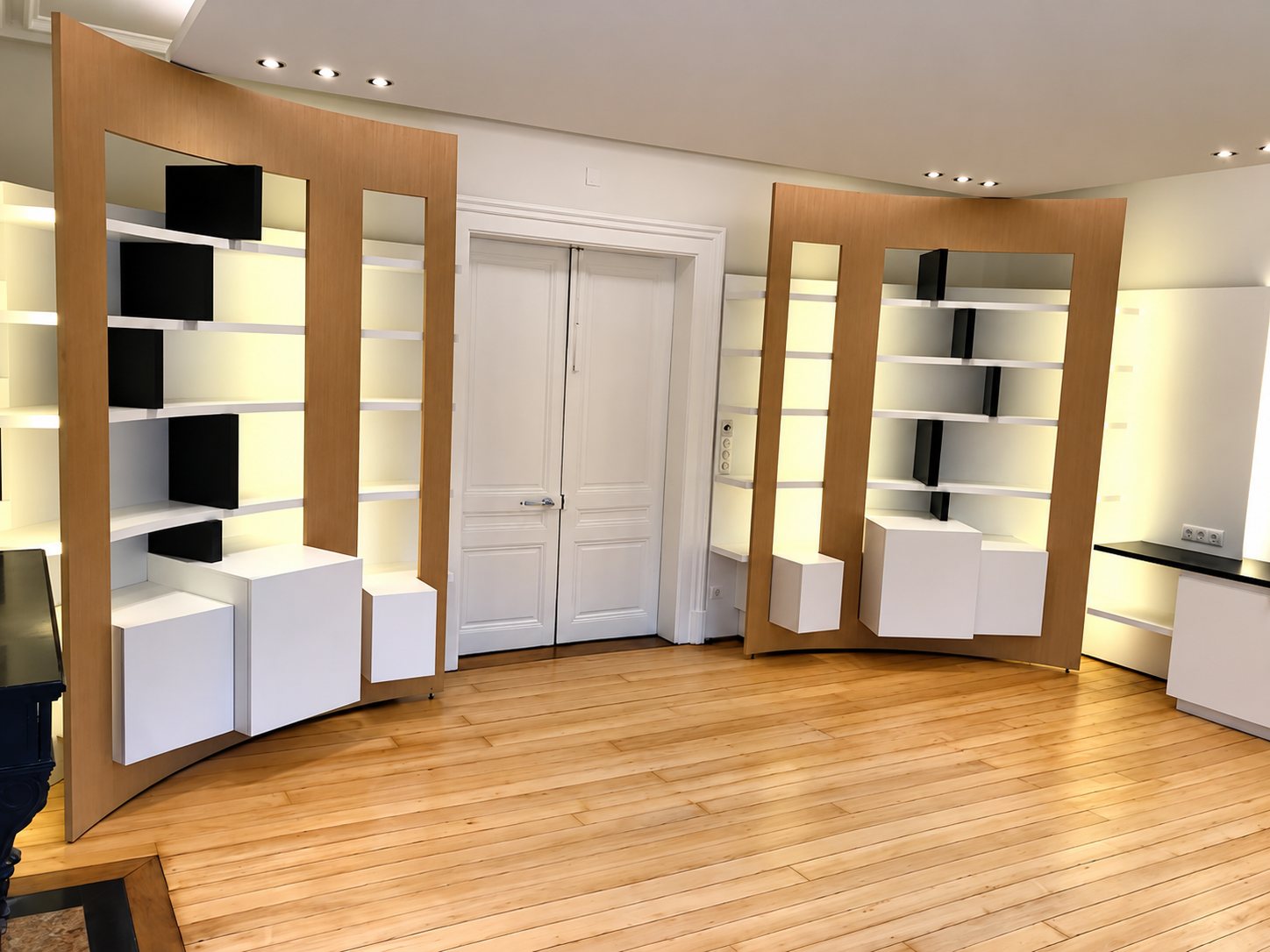 Deux grandes bibliothèques courbes en bois beige avec étagères blanches et éclairage intégré, de part et d'autre d'une porte double blanche dans une pièce au parquet clair.