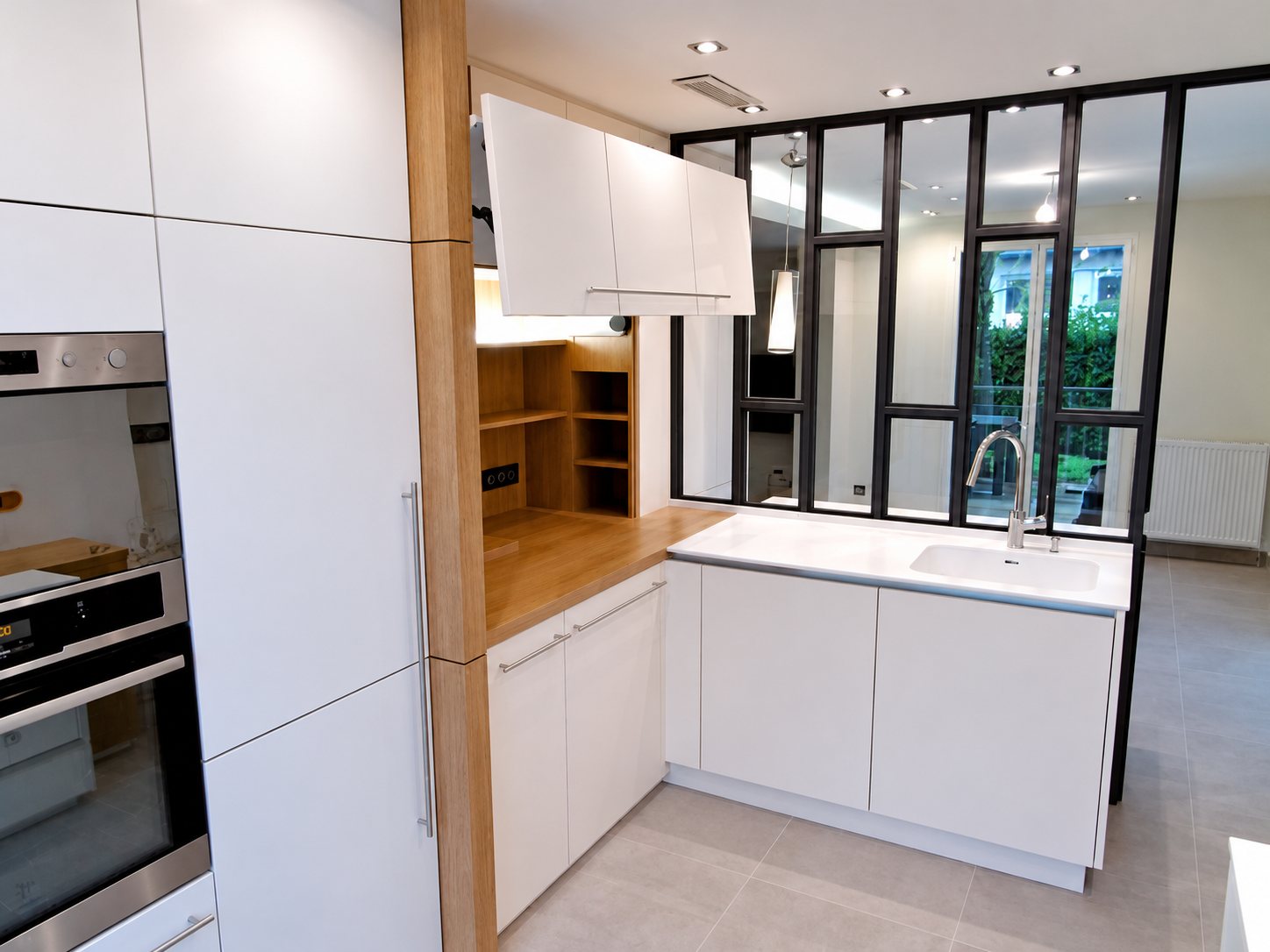Cuisine équipée aux façades blanches et aux éléments en bois clair, avec îlot à évier intégré et verrière d'atelier noire donnant sur la pièce voisine.