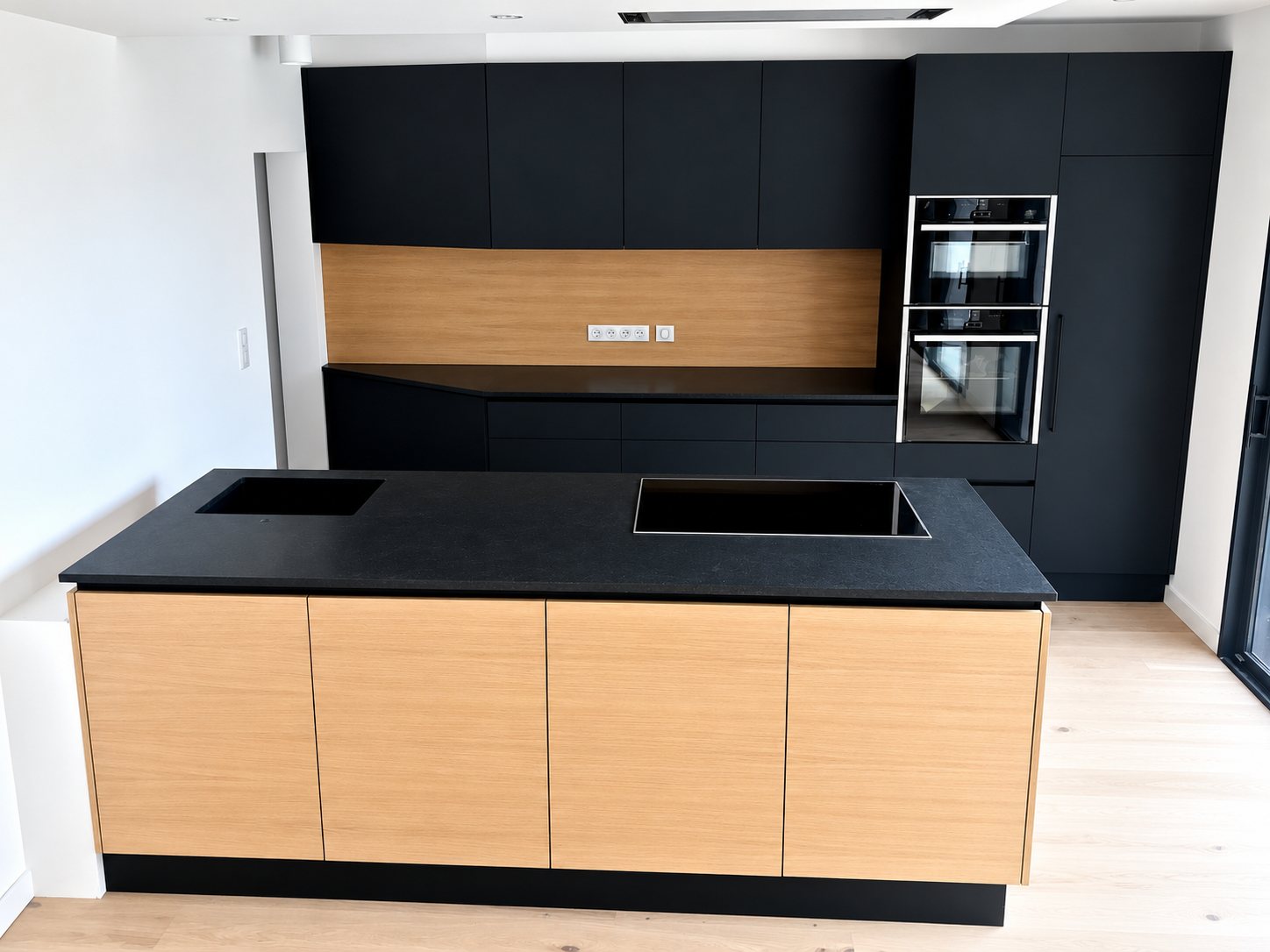 Cuisine contemporaine aux meubles noirs mats, crédence et façades d'îlot en bois clair, avec deux fours encastrés et plaque de cuisson sur l'îlot.