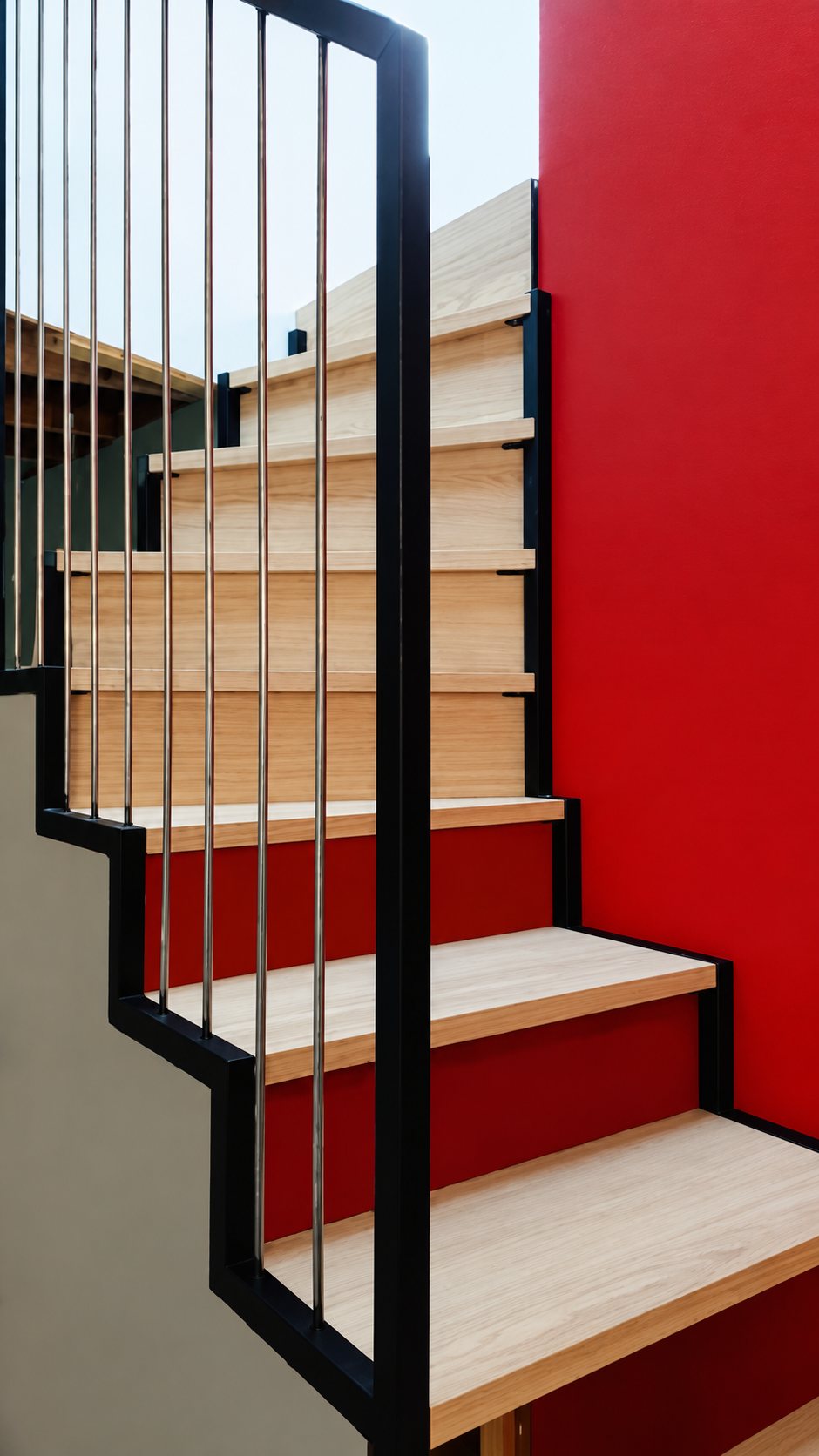 Escalier intérieur aux marches en bois clair, encadré par un garde-corps noir à tiges verticales et un mur rouge vif.