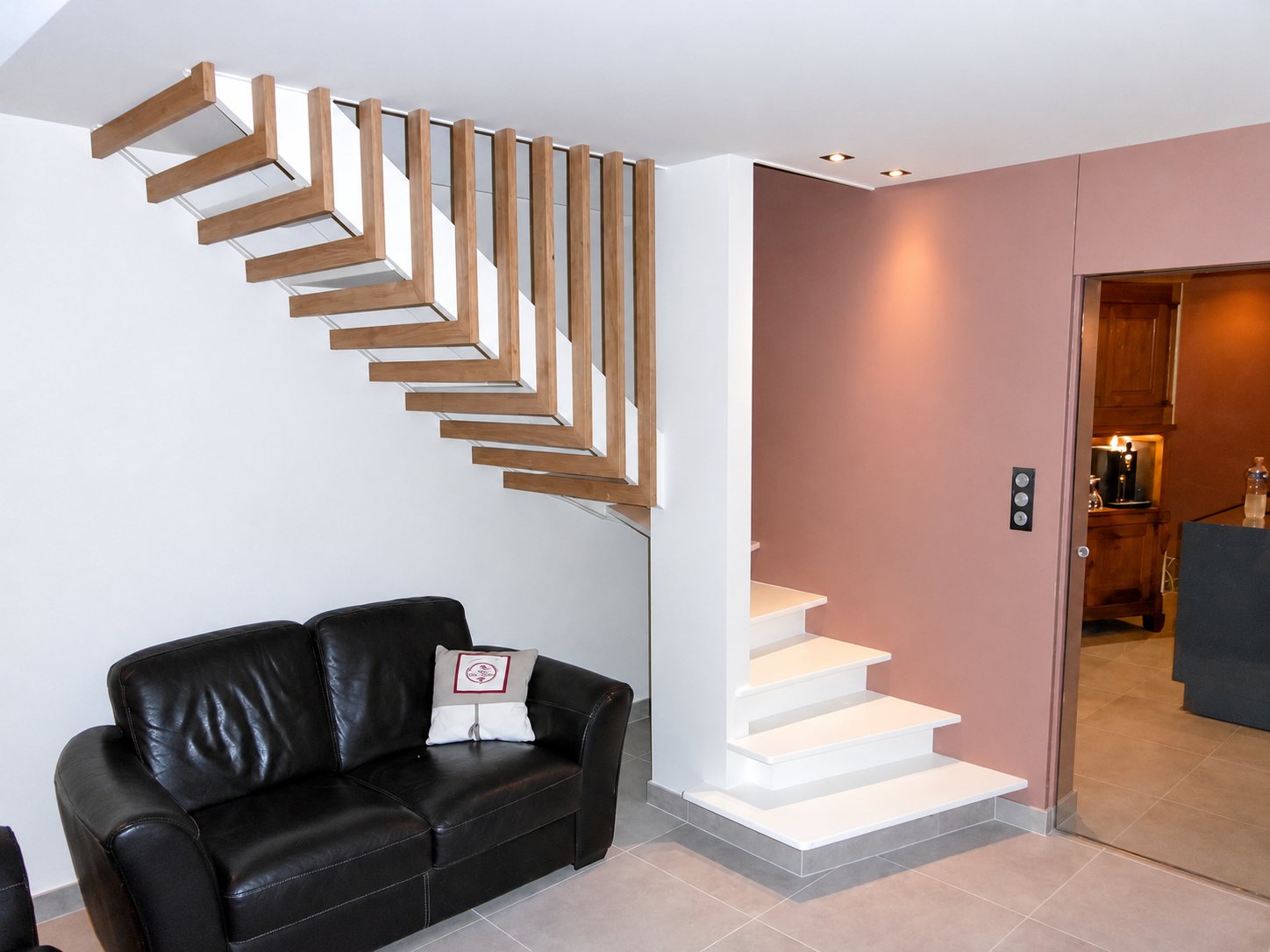 Un escalier contemporain tournant avec marches en bois clair suspendues et marches blanches, à côté d'un canapé en cuir noir dans un salon aux murs blancs et vieux rose.