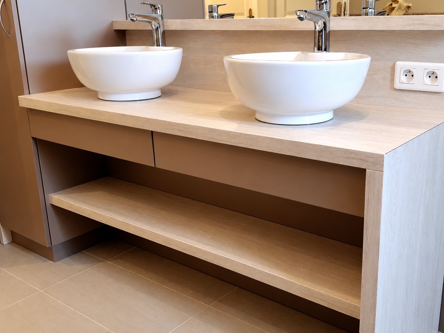 Meuble de salle de bain en bois clair équipé de deux vasques blanches à poser, de tiroirs et d'une étagère ouverte, installé dans une salle de bain.