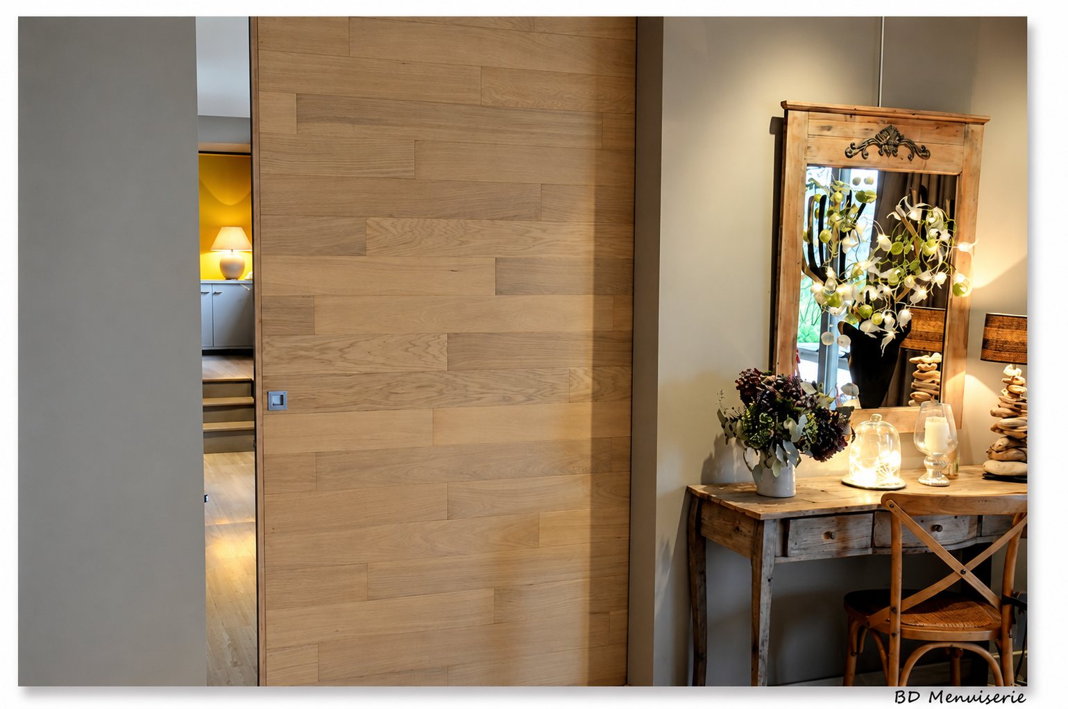 Une grande porte coulissante en lames de chêne clair ferme un couloir, à côté d'une console en bois surmontée d'un miroir ancien et de décorations florales.